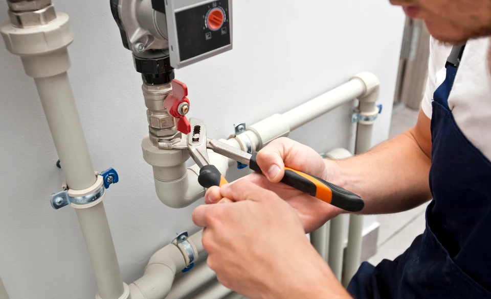 Plombier en combinaison bleue intervenant sur installation sanitaire intérieure, utilisant pince à main sur raccorderie